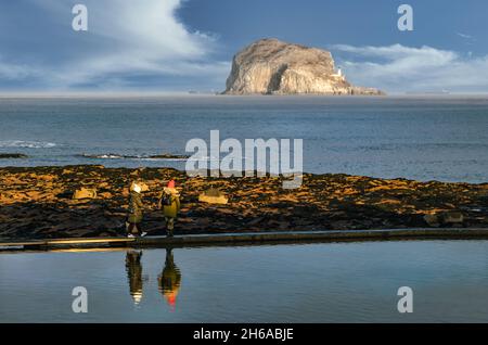 Zwei Personen, die auf einem ummauerten Meerwasserpool spazieren, spiegeln sich im stillen Wasser mit der Bass Rock Insel in der Ferne, North Berwick, Schottland, Großbritannien Stockfoto