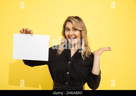 Junge schöne blonde Frau in schwarzem Kleid hält leeres Blatt für die Beschriftung auf gelbem Hintergrund in Händen zeigt mit der Hand, lächelt und lacht happ Stockfoto