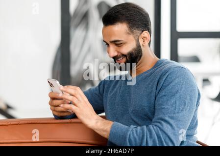 Fröhlicher Inder verbringt seine Freizeit damit, Nachrichten auf dem Smartphone zu scrollen, bärtiger Kerl hält Handy, schaut auf den Bildschirm, websurft, kauft, chattet in den sozialen Netzwerken, die auf der Couch liegen Stockfoto