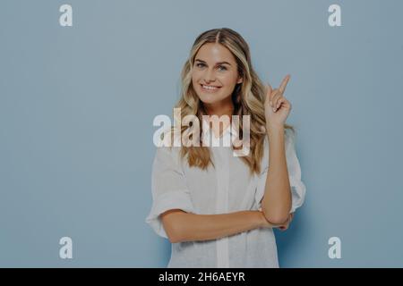 Fröhliche junge blonde Frau mit welligen langen Haaren, die mit dem Zeigefinger nach oben zeigt Stockfoto