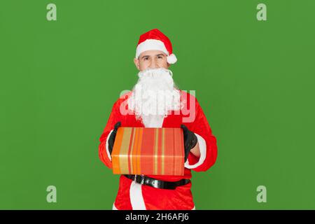 Ein erwachsener kaukasischer Mann, der als Weihnachtsmann gekleidet ist, liefert eine Geschenkbox nach vorne. Der Hintergrund ist grün. Stockfoto