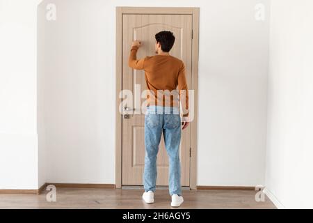 Rückansicht eines jungen, lässigen Mannes, der an die Holztür klopft, eines lässigen männlichen Besuchers, der im Eingang steht, Gast will herein, volle Körperlänge, Fr. Stockfoto
