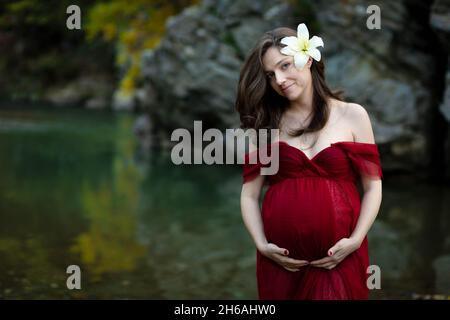 Junge, elegante, blonde Schwangere, die im Fluss in rotem Kleid, weißer Blume hinter ihrem Ohr steht Stockfoto