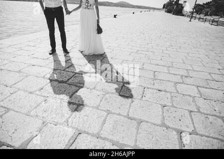 Braut und Bräutigam halten die Hände und stehen auf den Pflastersteinen am Meer. Schwarzweiß-Fotografie Stockfoto