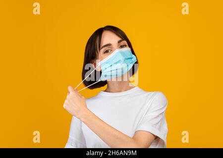 Ende der Epidemie. Junge Dame, die Schutzmaske auszieht und isoliert über gelbem Studiohintergrund steht. Schutz- und Gesundheitskonzept Stockfoto