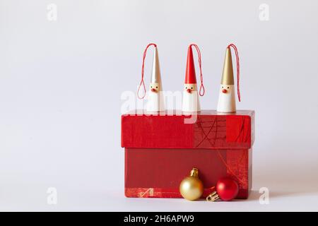 Drei Weihnachtsmänner-Figuren auf einer roten Geschenkbox, dekorative Weihnachtsbaumkugeln, weißer Hintergrund, Seitenansicht, Kopierraum Stockfoto