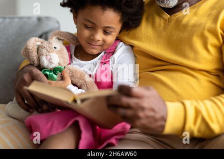 Lächelndes kleines afroamerikanisches Mädchen mit Spielzeug liest ein Buch mit einem älteren Großvater auf dem Sofa in der Inneneinrichtung, abgeschnitten, aus nächster Nähe. Studieren, Märchen togeth Stockfoto