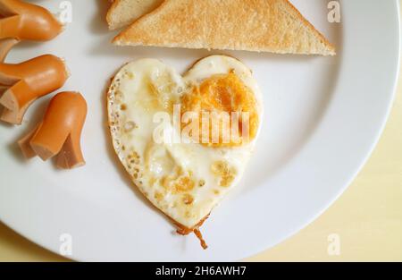 Köstliches Frühstück mit herzförmigen Spiegelei, Würstchen und Toast Stockfoto