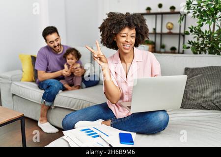 Mutter einer interracial Familie, die zu Hause mit einem Computer-Laptop arbeitet, lächelt mit einem glücklichen Gesicht, das die Kamera anwinkelt und mit den Fingern ein Siegeszeichen tut. Taub Stockfoto