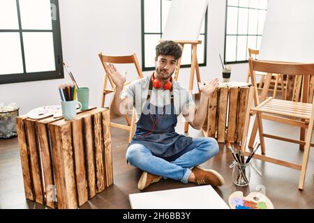 Junger hispanischer Mann sitzt im Kunststudio ahnungslos und verwirrt Ausdruck mit Armen und Händen angehoben. Zweifel Konzept. Stockfoto