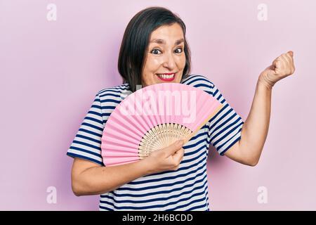 hispanische Frau mittleren Alters winkende Handventilator kühlende Luft im Sommer schreiend stolz, feiert Sieg und Erfolg sehr aufgeregt mit erhobenem Arm Stockfoto