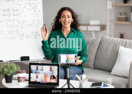 Weibliche Tutorin in Kopfhörern spricht, Videoanrufe online auf Handy-Bildschirm auf Stativ, mit pc zu Konferenz-Chat kommunizieren mit verschiedenen st Stockfoto