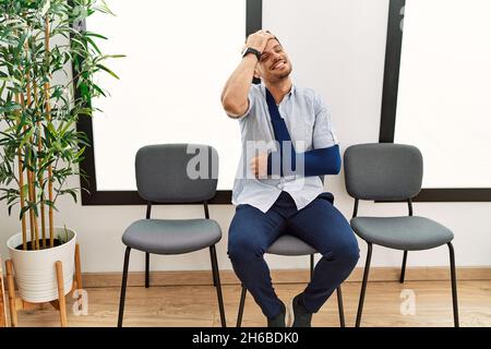 Hübscher junger Mann, der im Warteraum des Arztes sitzt und mit einer Armverletzung lächelt und selbstbewusst die Haare mit der Hand nach oben berührt, attraktiv und modisch posiert Stockfoto