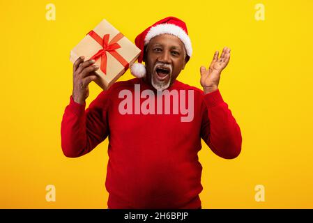 Aufgeregter älterer schwarzer Mann in rotem Pullover und Weihnachtsmannhut mit Geschenkbox, schreiend HO HO, mit großem Urlaubsverkauf oder Rabatt auf orange studi Stockfoto