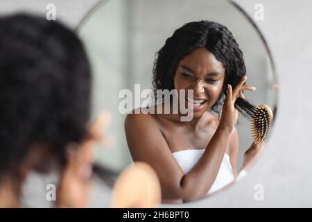 Irrende schwarze Dame mit buschigen Haaren, die vor dem Spiegel im Badezimmer stand und ihr Haar mit einem Holzpinsel kämmte, Schwierigkeiten hatte, Nahaufnahme. Hai Stockfoto