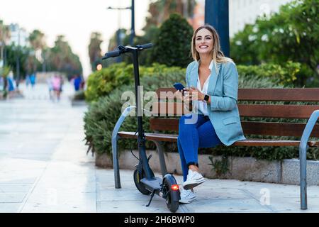 Stilvolle Geschäftsfrau mit Smartphone auf der Bank in der Nähe von Elektroroller Stockfoto