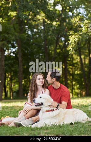 Vertikale Ganzkörperportrait von jungen Paar mit Hund genießen Picknick im Freien auf grünem Gras Stockfoto