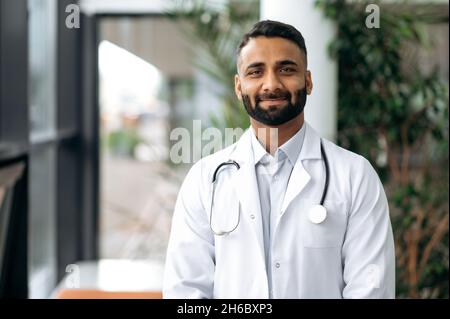 Porträt eines hübschen indischen Hausarztes in medizinischer Uniform und Stethoskop, der im Krankenhaus steht, die Kamera anschaut und freundlich lächelt Stockfoto