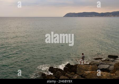 Einsamer Fischer, der an einem bewölkten Sommertag von einem Felsen an der Küste mit dem Vorgebirge von Capo Mele über den Meereshorizont fischt, Alassio, Savona Stockfoto