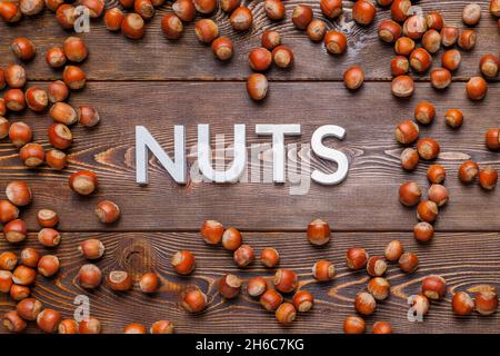 Das Wort Nüsse mit silbernen Buchstaben auf Holzbrett Hintergrund mit verstreuten Haselnüssen umgeben gelegt Stockfoto