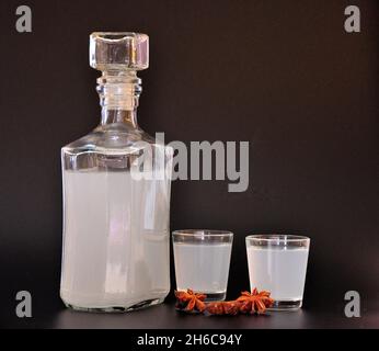 Anislikör, ein großes alkoholisches Getränk in einer Glasflasche und zwei Gläser auf schwarzem Hintergrund, neben den Sternen der getrockneten Gewürze. Nahaufnahme. Stockfoto