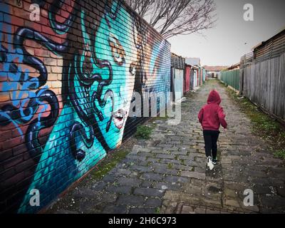 Ein Kind in einer roten Kapuzenjacke läuft im August 2021 in West Footscray, Melbourne, Australien, über einen graffierten Laneway. Stockfoto