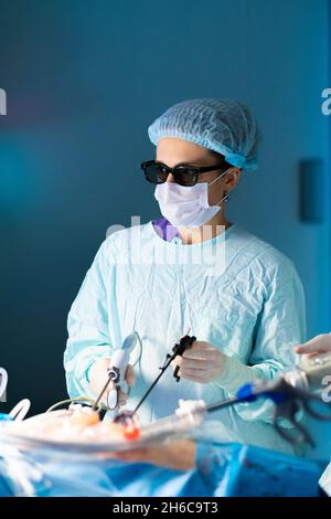 Eine Chirurgin operiert an einem Patienten. Selektiver Fokus. Der Chirurg hält laparoskopische Instrumente. Minimalinvasive Chirurgie. Arzt in OP-Uniform, Maske und 3D-Brille. Stockfoto