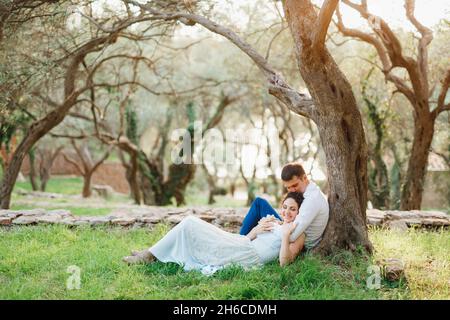 Kotor, Montenegro - 21.06.17: Sitzender Mann umarmt eine Frau, die neben ihm auf grünem Gras liegt, und küsst sie auf der Krone neben einem Baum in einem Olivenhain Stockfoto