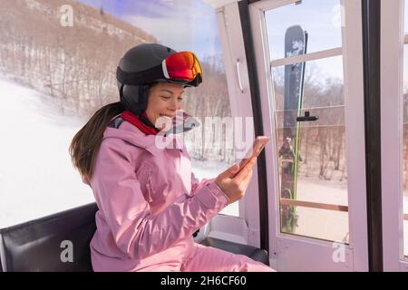 Skiurlaub - Frau Skifahrerin mit Telefon-App in Gondelbahn. Mädchen lächelnd Blick auf mobile Smartphone trägt Skibekleidung, Helm und Brille Stockfoto