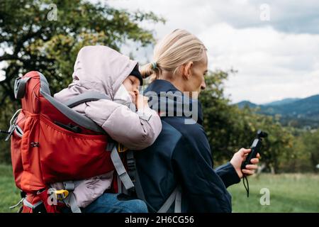 Vater fotografiert beim Wandern mit Kind im Rucksack. Vlogger-Mann, der mit einem erlafften Kind im Rucksack in den kalten Bergen läuft. Aktive Zeit mit Stockfoto