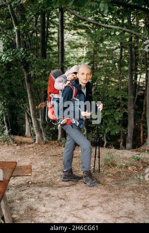 Mann, der mit einem verklenkten Kleinkind in den Berg wandert. Kind sitzt im Rucksack auf dem Rücken des Vaters. Aktives Familienabenteuer im Wald mit müdem Mädchen. Kalt Stockfoto