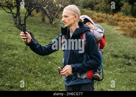 Vater fotografiert beim Wandern mit Kind im Rucksack. Vlogger-Mann, der mit einem erlafften Kind im Rucksack in den kalten Bergen läuft. Aktive Zeit mit Stockfoto