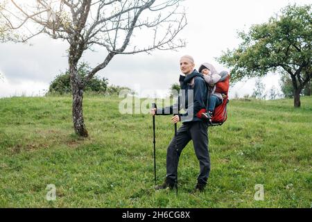 Mann, der mit einem verklenkten Kleinkind in den Berg wandert. Kind sitzt im Rucksack auf dem Rücken des Vaters. Aktives Familienabenteuer im Wald mit müdem Mädchen. Kalt Stockfoto