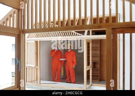 Kontraktoren in orangefarbener Uniform und Harthüten diskutieren Holzhausrahmen Stockfoto