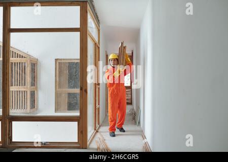 Starke seriöse Baumeister in orange Uniform hält schwere Holzgitter und Rahmen, die im Haus zu Bett installiert haben Stockfoto