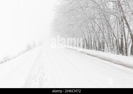 Winterlicher Schneefall auf der Straße. Eine leere Landstraße ohne Autos, bedeckt mit Schneeverwehungen. Reisen Sie bei schlechtem Wetter. Winter wolkige Landschaft mit einer Landstraße. T Stockfoto