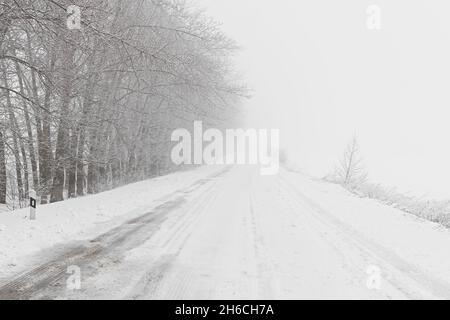 Winterlicher Schneefall auf der Straße. Eine leere Landstraße ohne Autos, bedeckt mit Schneeverwehungen. Reisen Sie bei schlechtem Wetter. Winter wolkige Landschaft mit einer Landstraße. T Stockfoto