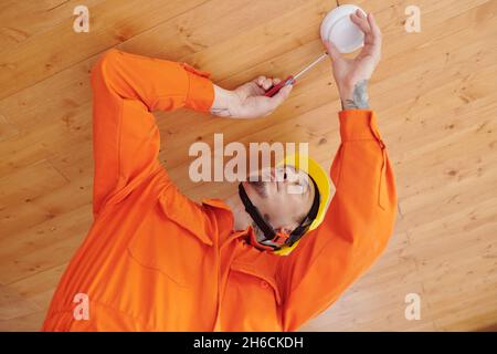 Techniker mit Schraubendreher, der Rauchmelder an der Holzdecke installiert Stockfoto