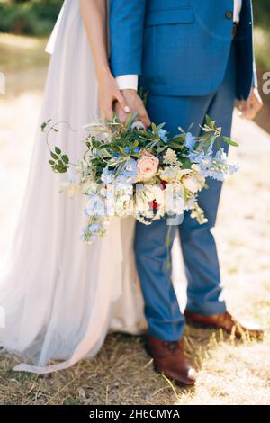 Bräutigam in einem blauen Anzug hält einen Blumenstrauß in der Hand. Braut in einem weißen Kleid umarmt ihn von hinten. Nahaufnahme Stockfoto