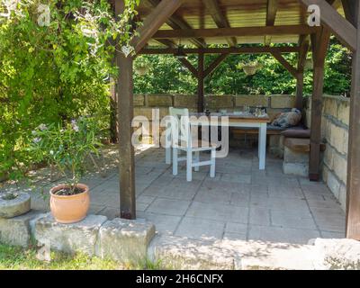 Gemütliche braune tiber Holzpavillon, Pergola mit weißem Tisch, Stühlen und Bank mit Kissen und Decke, umgeben von Sandsteinwand und Kletterpflanzen Stockfoto