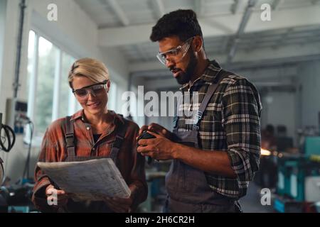 Porträt junger birazialer Industriekollegen, die in Innenräumen in einer Metallwerkstatt arbeiten, lächelnd. Stockfoto
