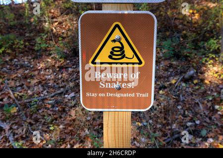 Ein Warnschild zur Vorsicht vor Schlangen auf einem Wanderweg, auf dem die Menschen darauf hingewiesen werden, auf einem ausgewiesenen Wanderweg zu bleiben. Stockfoto