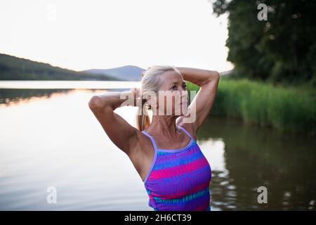 Porträt einer aktiven älteren Schwimmerin, die im Freien im See steht und sich ausdehnt. Stockfoto