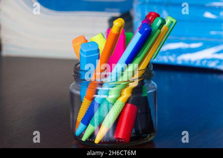 Mehrfarbige Stifte und Marker in einem Standfuß auf dem Hintergrund des Stapels von Notizbüchern. Eine Packung blauer Schulnotizbücher auf dem Tisch im Klassenzimmer. Stapel Stockfoto