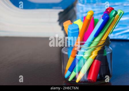 Mehrfarbige Stifte und Marker in einem Standfuß auf dem Hintergrund des Stapels von Notizbüchern. Eine Packung blauer Schulnotizbücher auf dem Tisch im Klassenzimmer. Stapel Stockfoto