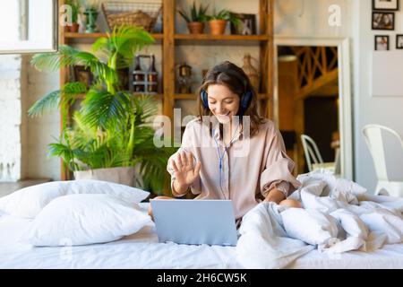 35-jährige Frau während eines Online-Videoanrufs - Sie sitzt morgens auf dem Bett vor einem Laptop-Monitor. Modernes Technologiekonzept. Stockfoto
