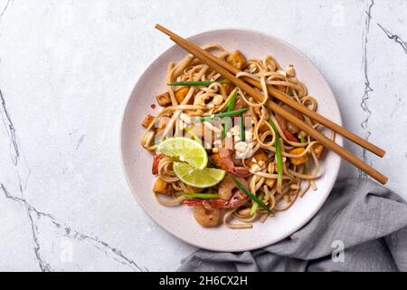 Draufsicht auf Teller mit asiatischen udon-Nudeln, würzigen Garnelen, gebratenem Tofu-Käse, Erdnüssen, Limette und grünen Zwiebeln auf weißem Marmorboden Stockfoto