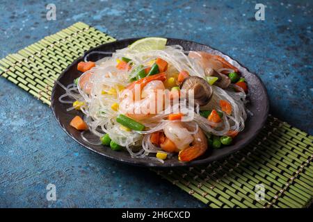 Schwarzer Teller mit japanischen Bohnen- oder Glasnudeln, leckeren Garnelen, Shiitake-Pilzen und Gemüse auf blauem Betongrund Stockfoto