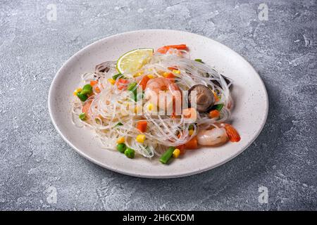 Teller mit japanischen Bohnen- oder Glasnudeln, leckeren Garnelen, Shiitake-Pilzen und Gemüse auf grauem Betongrund Stockfoto