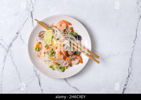 Draufsicht auf Teller mit japanischen Bohnen- oder Glasnudeln, leckeren Garnelen, Shiitake-Pilzen und Gemüse auf weißem Marmorboden Stockfoto